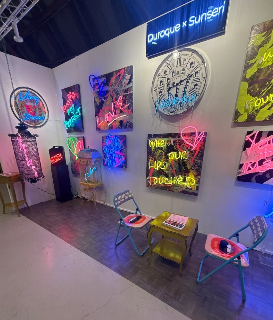 Interior view of Missy Sunseri's Barker Hangar exhibition booth showing real glass neon artworks and mixed-media paintings on white walls, with seating and a Duroque x Sunseri sign overhead